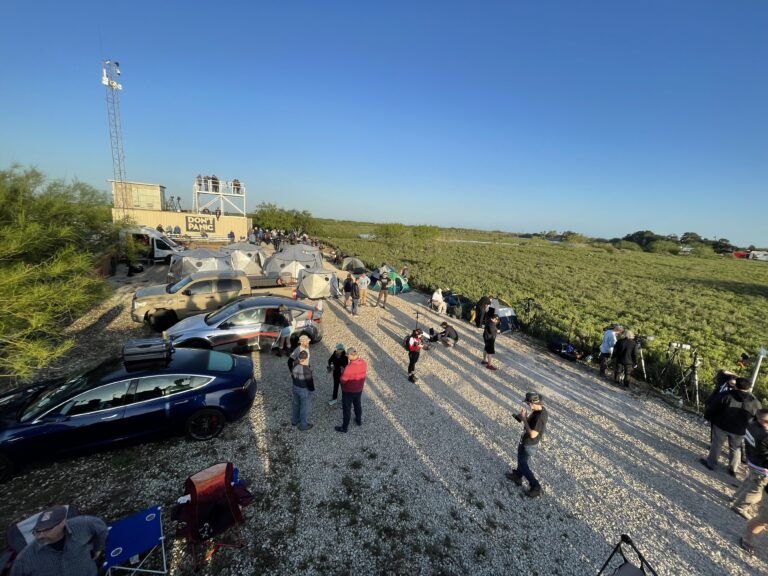 Launch-day crowd and cars lined up along the gravel viewing area at the Rocket Ranch Outpost, with the viewing tower and dome tents behind