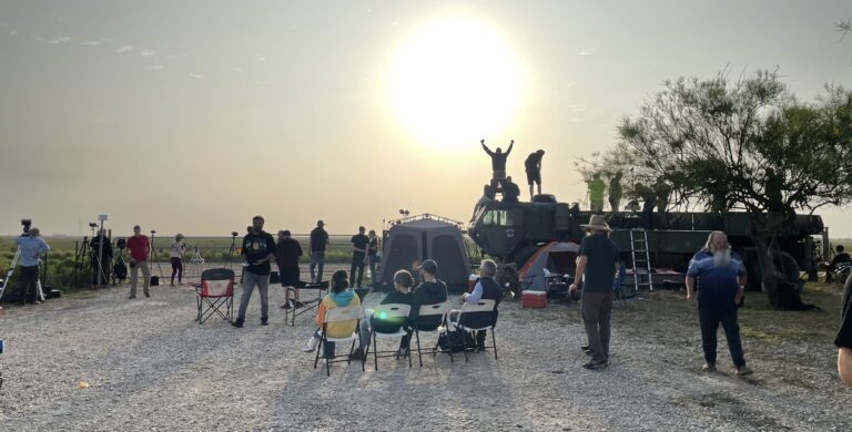 Guests in camp chairs at the Rocket Ranch Outpost with the sun low on the horizon and spectators on top of a military truck